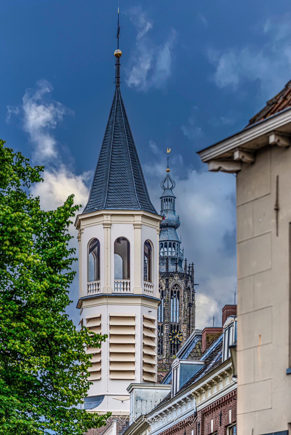 Een historische torenspits met de Onze Lieve Vrouwetoren van Amersfoort erachter, tegen een dramatische bewolkte lucht.