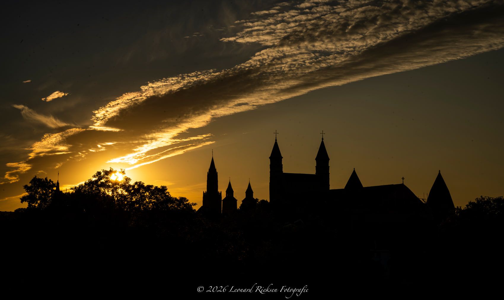 De torens en daken van de Maastrichtse binnenstad als donker silhouet tegen een oranje lucht met langgerekte wolkenbanen, kort na zonsondergang.