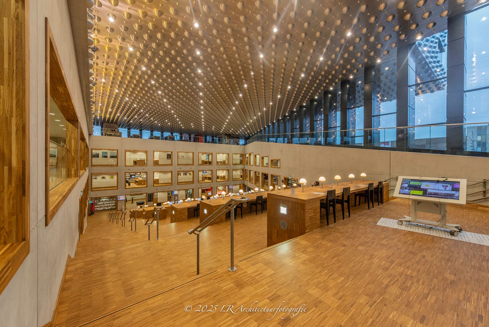 De leeszaal van het Eemhuis in Amersfoort met een golvend, verlicht plafond boven warme houten leestafels.